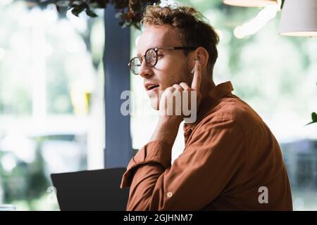 young freelancer in glasses adjusting earphones near laptop with blank screen in cafe Stock Photo