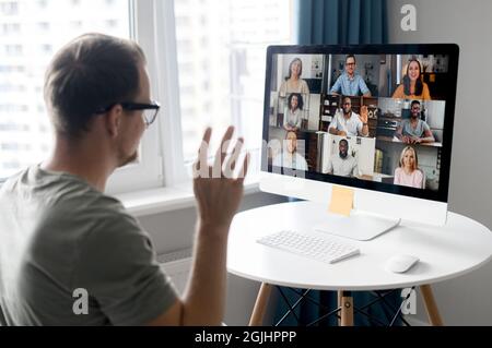 Attentive young hipster man sitting at table with PC, working remotely from home, on team meeting online with coworkers, looking at screen with people profiles and waving hand Stock Photo