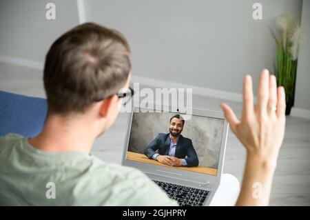 Young guy is using computer app for video connection on the laptop, talking online with male Indian friend, colleague or tutor, man is waving hand, greeting ethnic tutor or teacher, video call Stock Photo