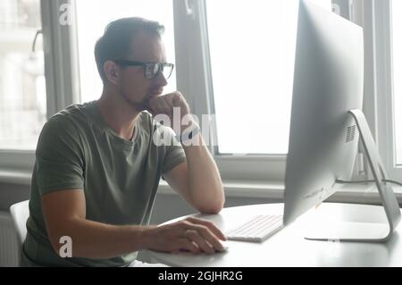 Serious focused determined young bearded businessman or freelancer in glasses and casual clothes sitting at kitchen table, using trendy PC, working on project, researching, writing email to client Stock Photo