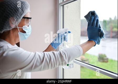 Mature woman in protective medical suit and respirator mask desinfecting furniture and cleaning window glass at hospital Stock Photo