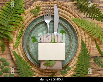 Blank paper card on table setting decorated with fern leaves on wooden table top view. Tropical mock-up scene with place card flat lay Stock Photo
