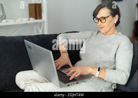 An intelligent mature woman wearing stylish glasses using a laptop working from home in relaxed atmosphere, elderly female concentrated types on the keyboard lying on the couch Stock Photo