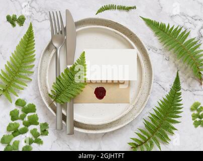 Blank paper card and sealed envelope on table setting decorated with fern leaves on white marble table top view. Tropical mock-up scene with place car Stock Photo