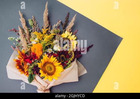 Fall bouquet of yellow red orange flowers wrapped in paper and arranged on grey and yellow background. Sunflowers, amaranth, daisies with zinnias and Stock Photo