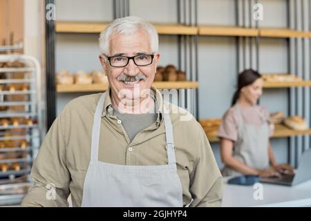 Portrait of smiling mature man in apron and eyeglasses standing against display with fresh bread in bakery house Stock Photo
