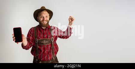 Young bearded man, waiter in traditional Bavarian costume using phone isolated over white studio background. Celebration, oktoberfest, festival Stock Photo