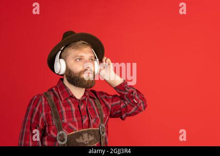 Comic portrait of bearded man, waiter in hat and traditional Bavarian costume isolated over red studio background. Oktoberfest, fest, holidays concept Stock Photo