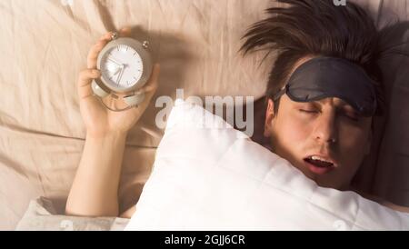 Hands of a young man from under the blankets hold an alarm clock in gray.  Stock Photo