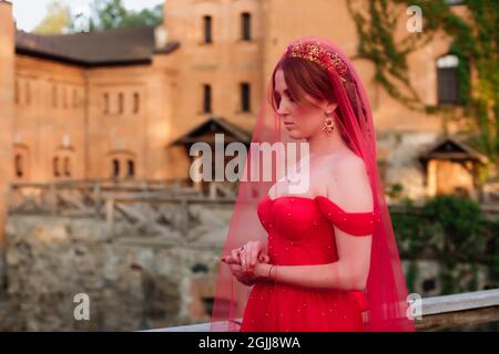 Portrait of attractive redhead tattooed woman in long red dress, diadema and red veil on blurred medieval castle background Stock Photo