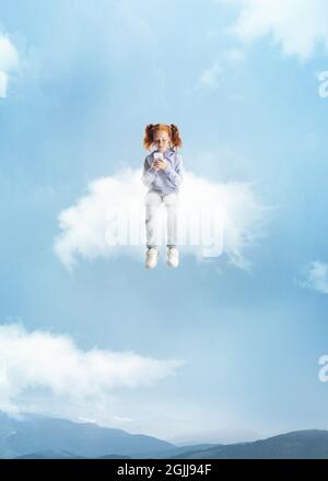 Conceptual collage with little beautiful girl sitting on white cloud using phone and flying at sky, outdoors. Concept of childhood, happiness, dreams Stock Photo