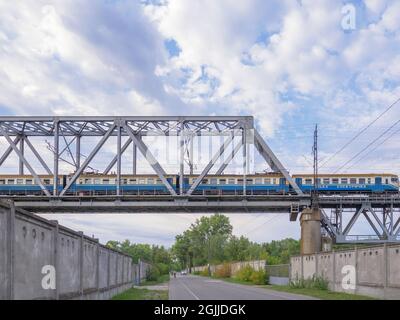 Suburban commute train going over the railroad bridge above the road with bike riders and a car in Kyiv (Kiev), Ukraine. Industrial cityscape concept. Stock Photo