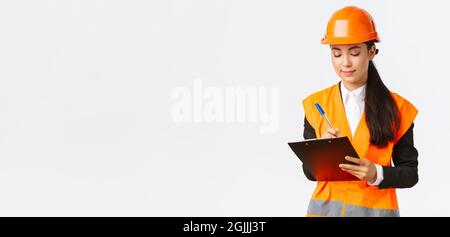 Pleased asian female construction engineer, architect taking notes in clipboard, writing down something during inspection at building area, wearing Stock Photo