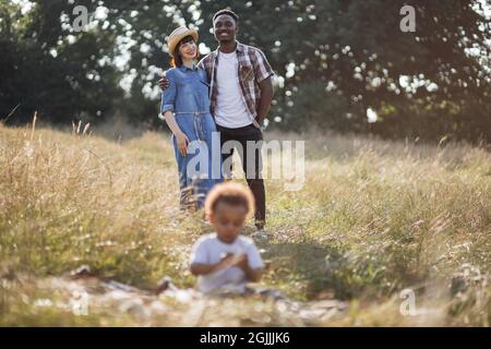 Blur foreground of little african boy sitting on grass and playing while his multiracial parents standing behind, hugging, smiling and looking at him. Happiness from parenthood. Stock Photo