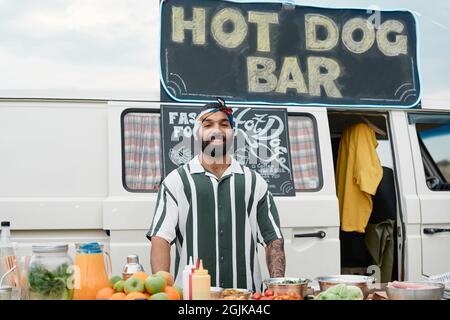 Portrait of young bearded man smiling at camera while selling food outdoors with van in the background Stock Photo