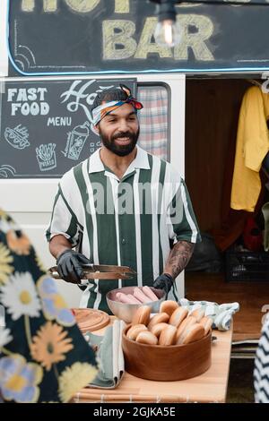 Young man in protective gloves cooking takeaway food while standing at the table with van in the background Stock Photo