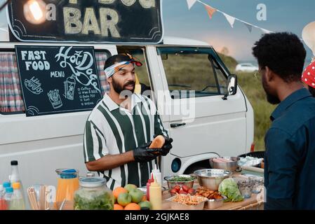 Young man standing at the table outdoors and cooking hot dogs with van in the background Stock Photo
