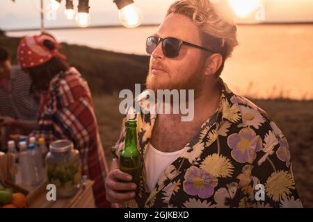 Young bearded man in sunglasses drinking beer with his friends at the party on the beach Stock Photo