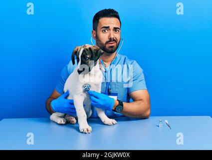 Handsome hispanic veterinary man with beard checking dog health using stethoscope clueless and confused expression. doubt concept. Stock Photo