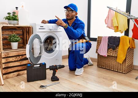 Young indian technician working on washing machine pointing with finger surprised ahead, open mouth amazed expression, something on the front Stock Photo