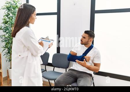 Youg latin injuried man wearing sling talking with the doctor at clinic waiting room. Stock Photo