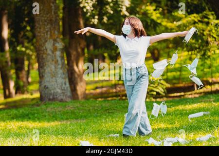 Full-length photo of a young woman wearing a facial medical mask holding hands by the sides and throwing medical mask around her outdoors. Stock Photo