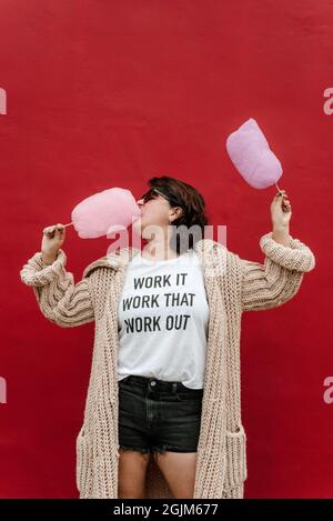 A woman bites into a pink cotton candy in profile. He holds another cotton in his other hand, he wears sunglasses, a long knitted sweater, and short p Stock Photo