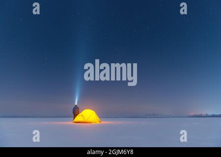 Tourist with flashlight near yellow tent lighted from the inside against the backdrop of incredible starry sky. Amazing night landscape. Tourists camp in snowy field. Travel concept Stock Photo