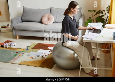 Serious young pregnant woman sitting on fitball in living room with mess on carpet and using laptop while working at home Stock Photo