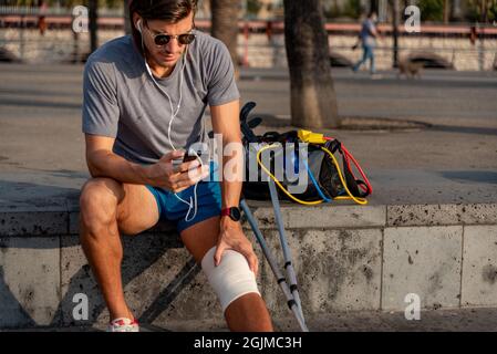 Young latin man with crutches using mobile phone after doing exercise Stock Photo