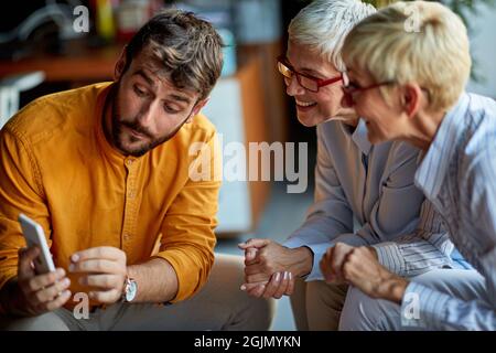A young male colleague is showing smartphone content to two older business women of similar appearance during a break in a friendly atmosphere at work Stock Photo