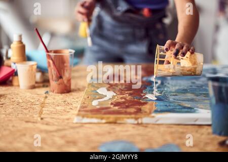 A young female artist is pouring the paint over a new painting she is working on in a pleasant atmosphere in the studio. Art, painting, studio Stock Photo