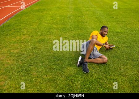 Happy young adult man laying on the grass, using smartphone, browsing internet. Joyful african american male relaxing, taking a break after workout Stock Photo