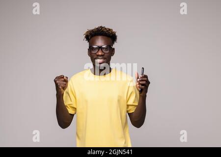 Excited african American millennial male in glasses isolated on grey studio background hold smartphone happy wining online, overjoyed black man look a Stock Photo