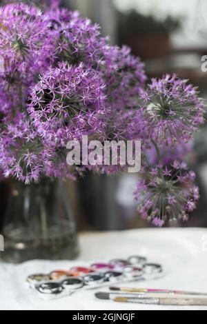 bouquet of blooming decorative onions of violet-lilac color close-up selective focus in a glass vase stands on the table next to brushes and watercolo Stock Photo