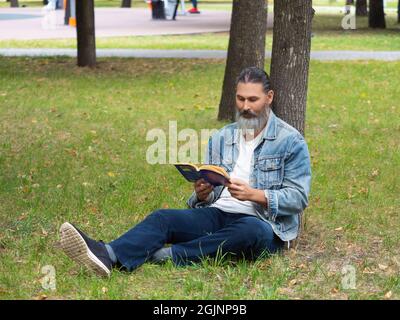 Bearded middle aged man reading the book in the park. He sitting on the grass in the park under the tree. The digital detox concept. Stock Photo