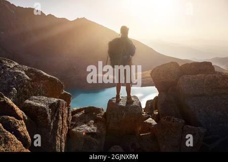 Silhouette of Man Hiker tourist man with backpack standing on the rock against beautiful sunset at lake in the mountain valley. Outdoor and trekking c Stock Photo