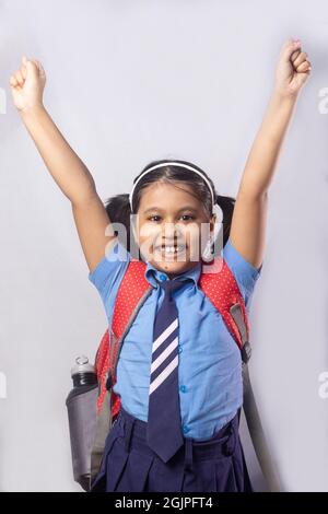 A happy cheerful smiling Indian girl child student in blue school uniform with red bag on grey background Stock Photo