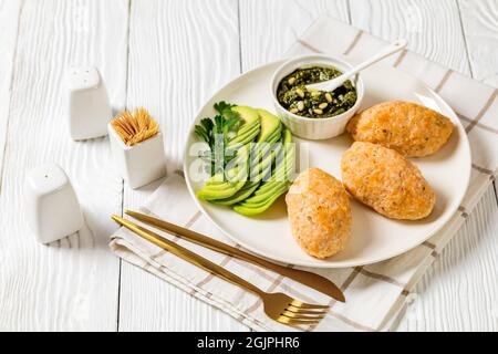 Baked in oven white fish patties or cakes served with green sauce pesto and sliced avocado on a white plate on a wooden table with golden cutlery Stock Photo