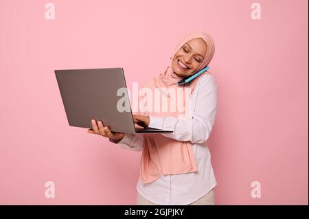 Successful Muslim Arab gorgeous woman in hijab, dressed business smart casual talks on mobile phone , smiles looking at the monitor screen while typin Stock Photo