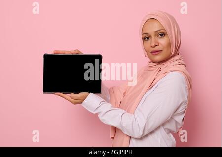 Confident Muslim Arab beautiful woman with covered head in hijab holds horizontally a digital tablet and shows its blank screen looking at the camera, Stock Photo
