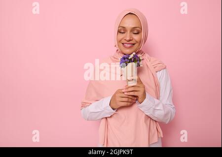 Portrait on pink background of beautiful Muslim Arab woman in hijab with closed eyes and charming smile enjoying the smelling of meadow flowers wrappe Stock Photo