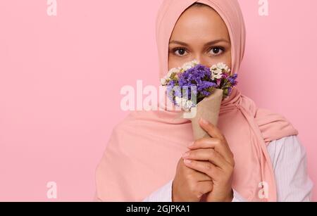 Close-up headshot portrait of young charming Arab Muslim woman in pink hijab with beautiful dark eyes, attractive gaze, looking at camera, covers half Stock Photo
