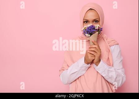 Confident portrait of young charming Arab Muslim woman in pink hijab with beautiful dark eyes, attractive gaze, looking at camera, covers half of her Stock Photo