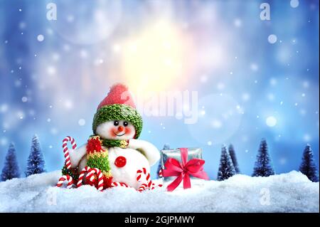 Cheerful snowman with gift box and candy cane on the snow. Christmas background. Holiday concept. Stock Photo