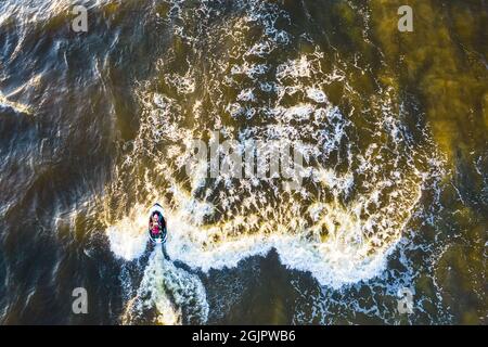 Aerial view of Young man rides on fast water scooter jumping on shallow waves and making golden splashes on sunset in blue water. Drone photo of tourist having fun riding a water bike on the sea Stock Photo