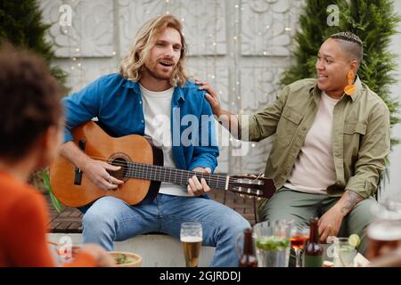 Portrait of handsome young man playing guitar and singing while enjoying outdoor party with friends, copy space Stock Photo