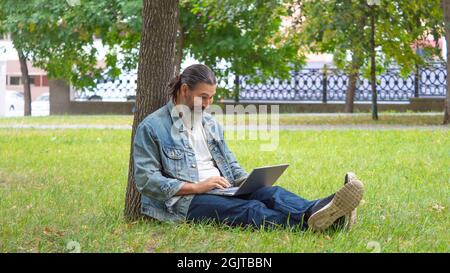Bearded middle aged man reading the book in the park. He sitting on the grass in the park under the tree. The digital detox concept. Format photo 16x9 Stock Photo