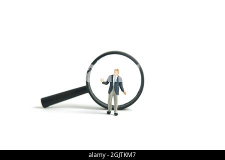 Miniature people toy figure photography. A shrugging businessman finding solution standing in front of magnifier glass. Isolated on white background. Stock Photo