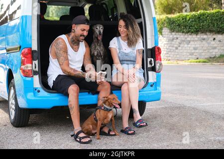 Young tattooed couple with their dogs using the phone in the back of the van. Stock Photo